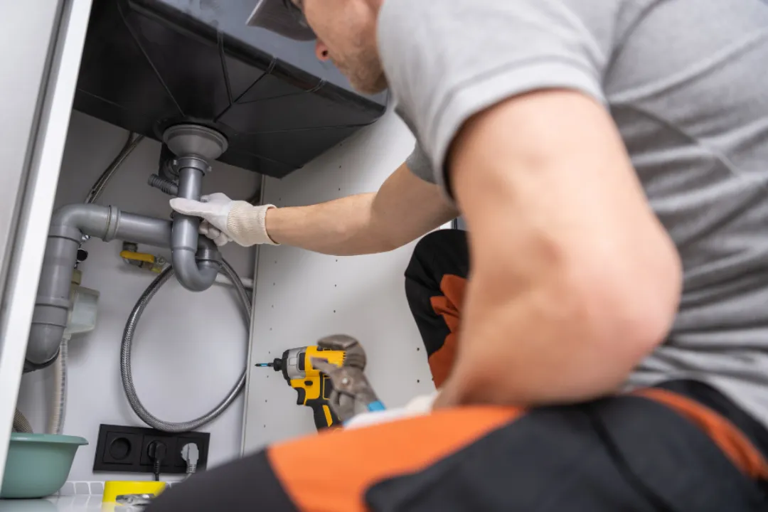 A man is engaged in fixing a sink, carefully handling a pipe while crouched under the countertop A man is engaged in fixing a sink, carefully handling a pipe while crouched under the countertop